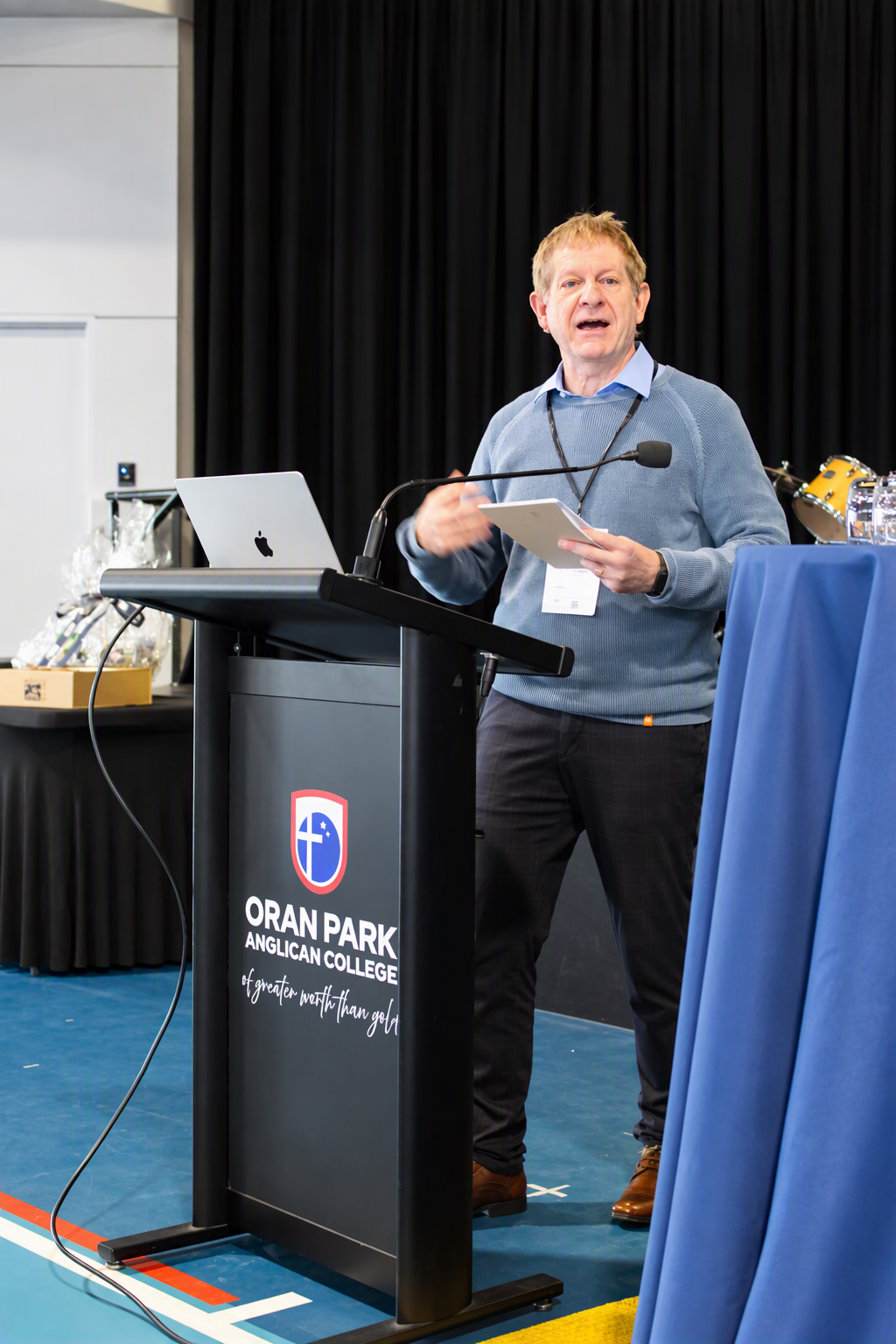 a man presents at the lectern in a light blue jumper. He is animated and holds a notepad.