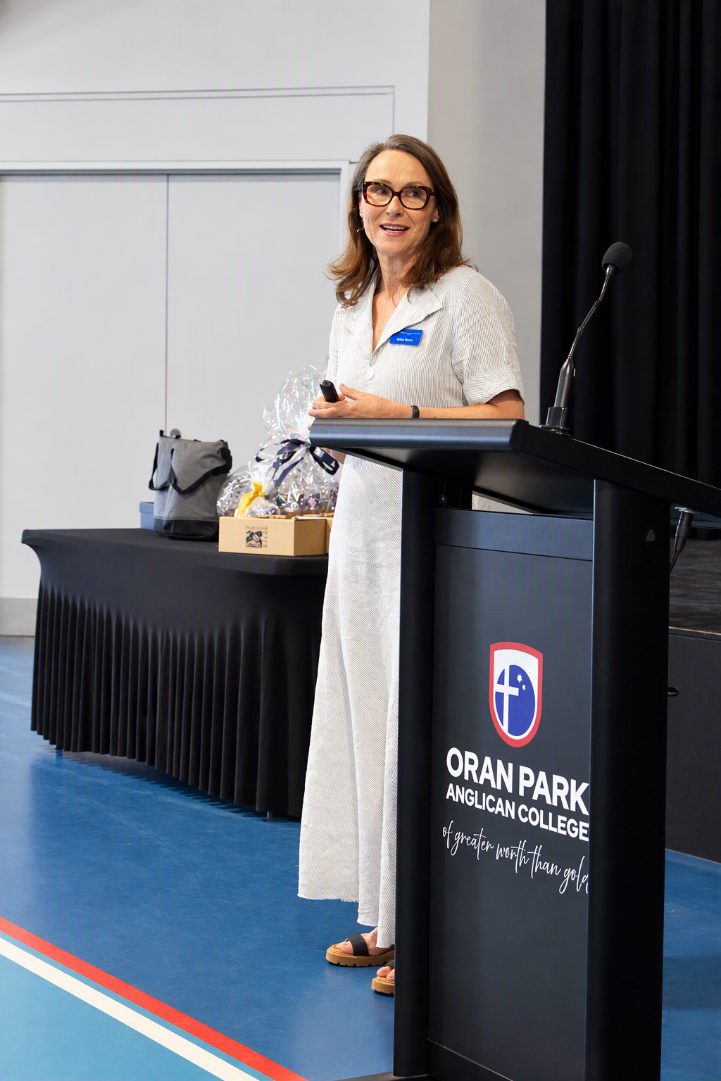 A women wearing a white dress and glasses stands to the side of the lectern presenting.