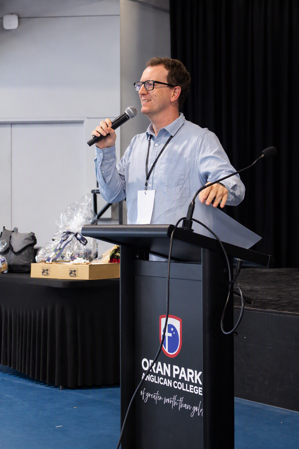 A man in glasses stands at the lectern holding a microphone. He is in a light blue dress shirt.